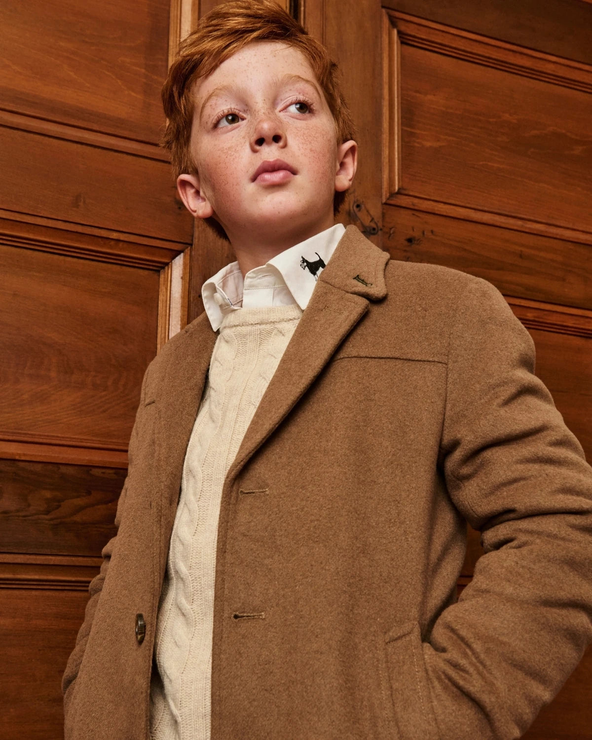 A boy wearing a camel wool coat, standing on a staircase, alternate view. Appaman Fall/Winter 2026 collection.