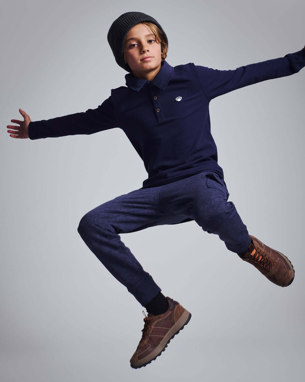 Boy in a navy blue Parker Polo jumping in stylish outfit with brown shoes and a beanie.