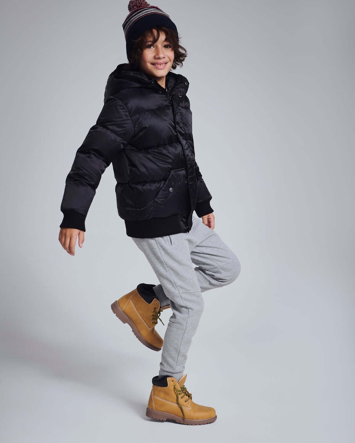 Boy wearing Novelty Grey Heather Sideline Sweatpants and a black puffer jacket, smiling with winter boots.