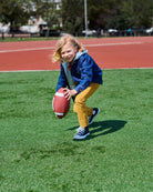 Child playing football on a grassy field wearing British Khaki Skinny Twill Pants and a blue jacket.
