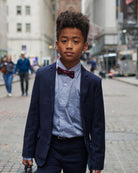 A stylish boy wearing a navy suit and a grey standard button down shirt with a bow tie in a city street.