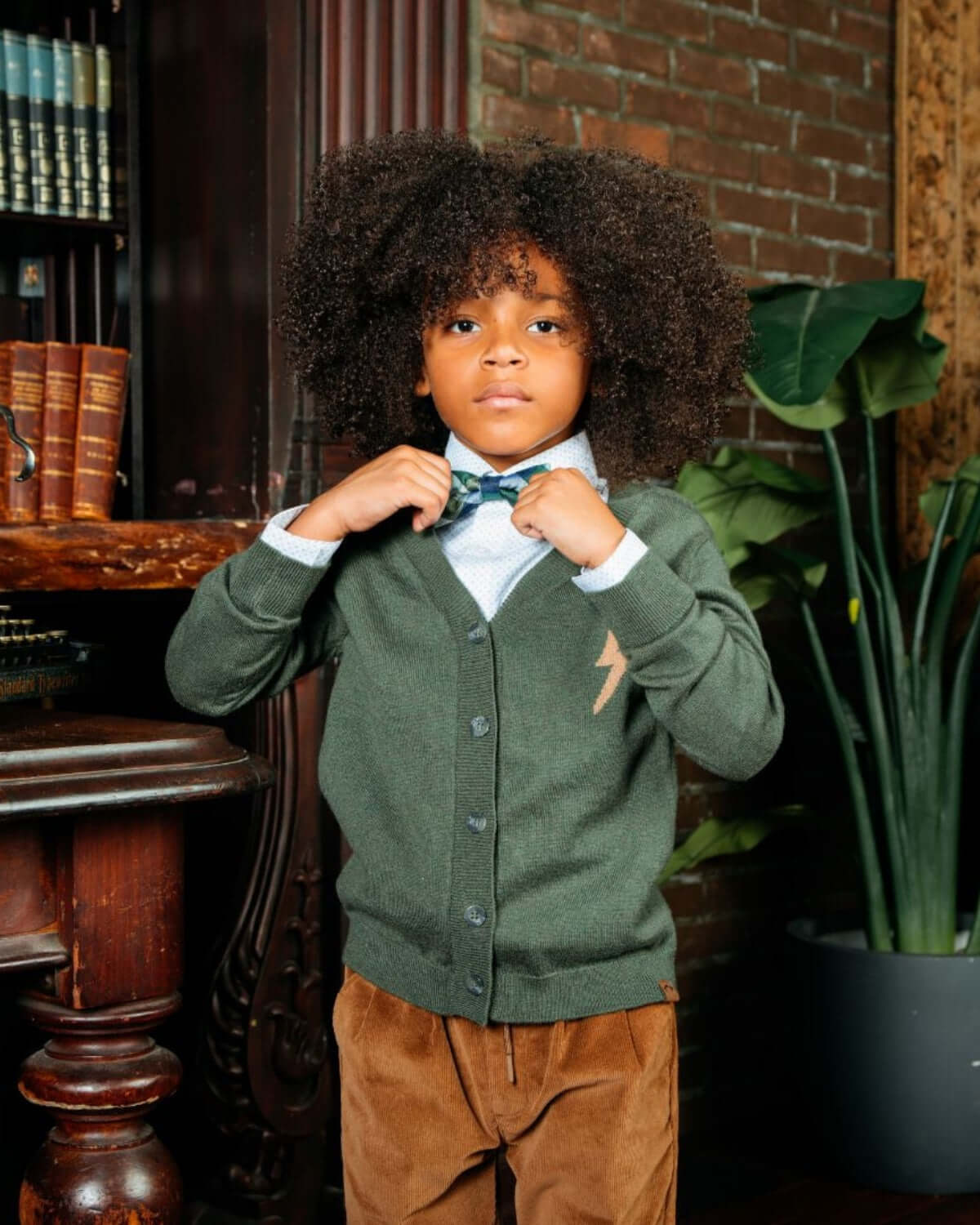 A young boy with curly hair wearing a green cardigan and bow tie, posing confidently in a stylish setting.