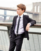A young boy in a suit with a Premium White Standard Button Down shirt, standing by a river with a cityscape in the background.