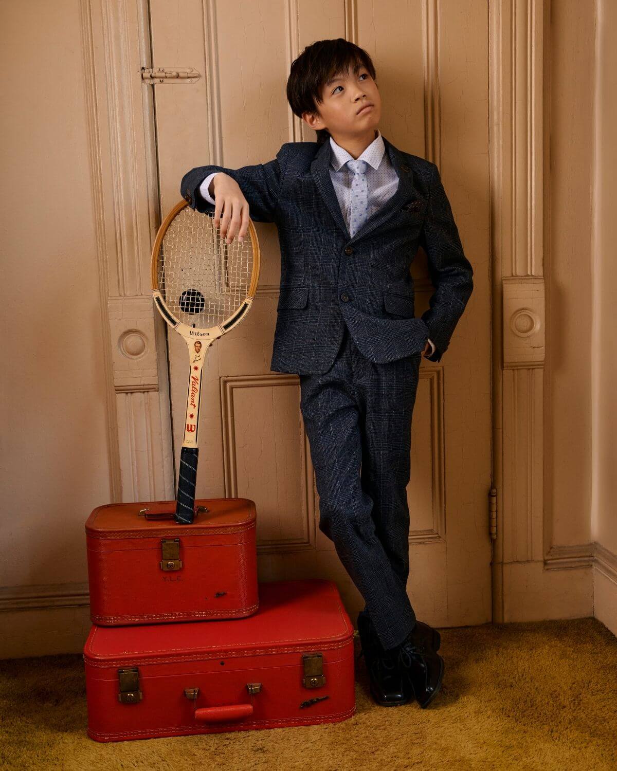A young boy in a Gotham Plaid Stretchy Mod Suit poses confidently with a tennis racket beside vintage suitcases.
