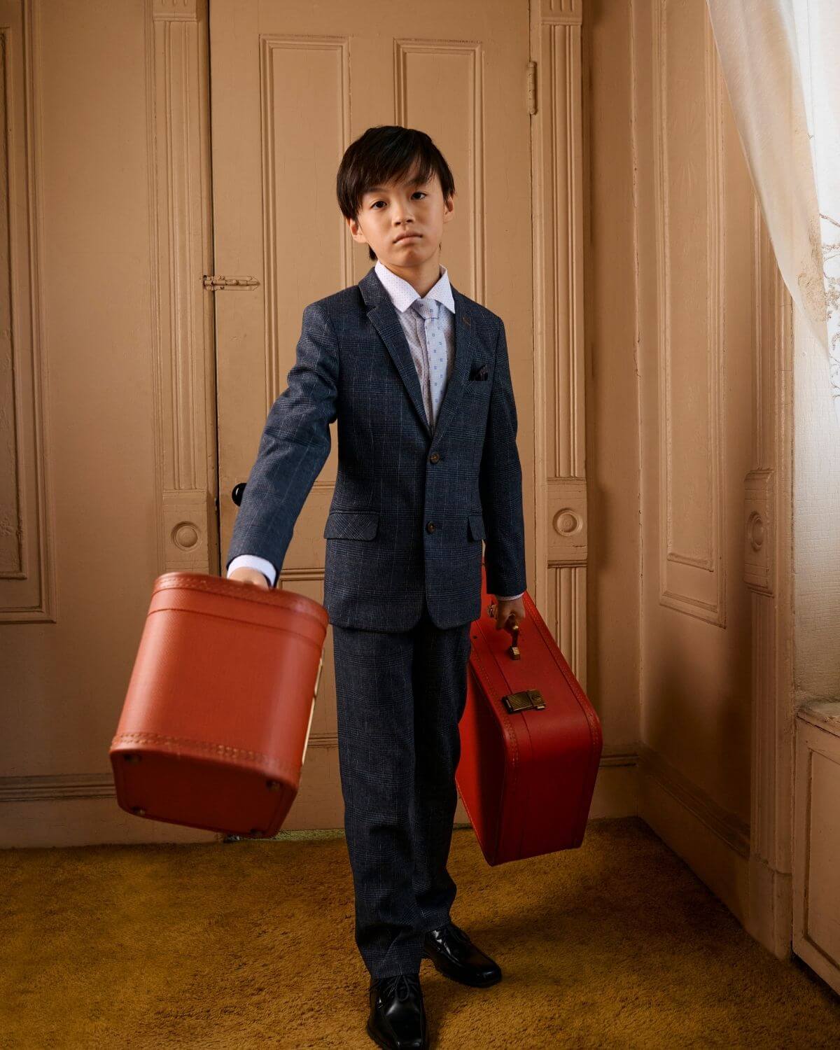 Boy in Gotham Plaid Stretchy Mod Suit holding two red suitcases, showcasing modern children's fashion.