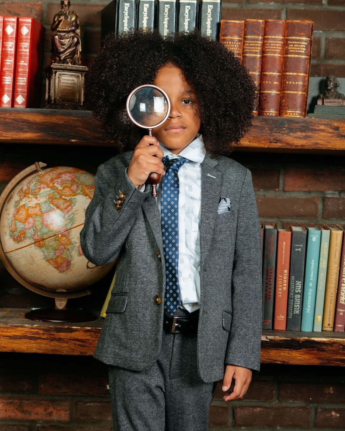 Boy wearing a Noir Micro Plaid Stretchy Mod Suit with a magnifying glass in a library setting.