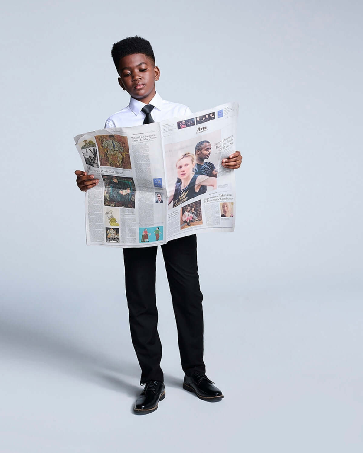 A young boy wearing a white shirt and black suit pants, reading a newspaper with focused attention.