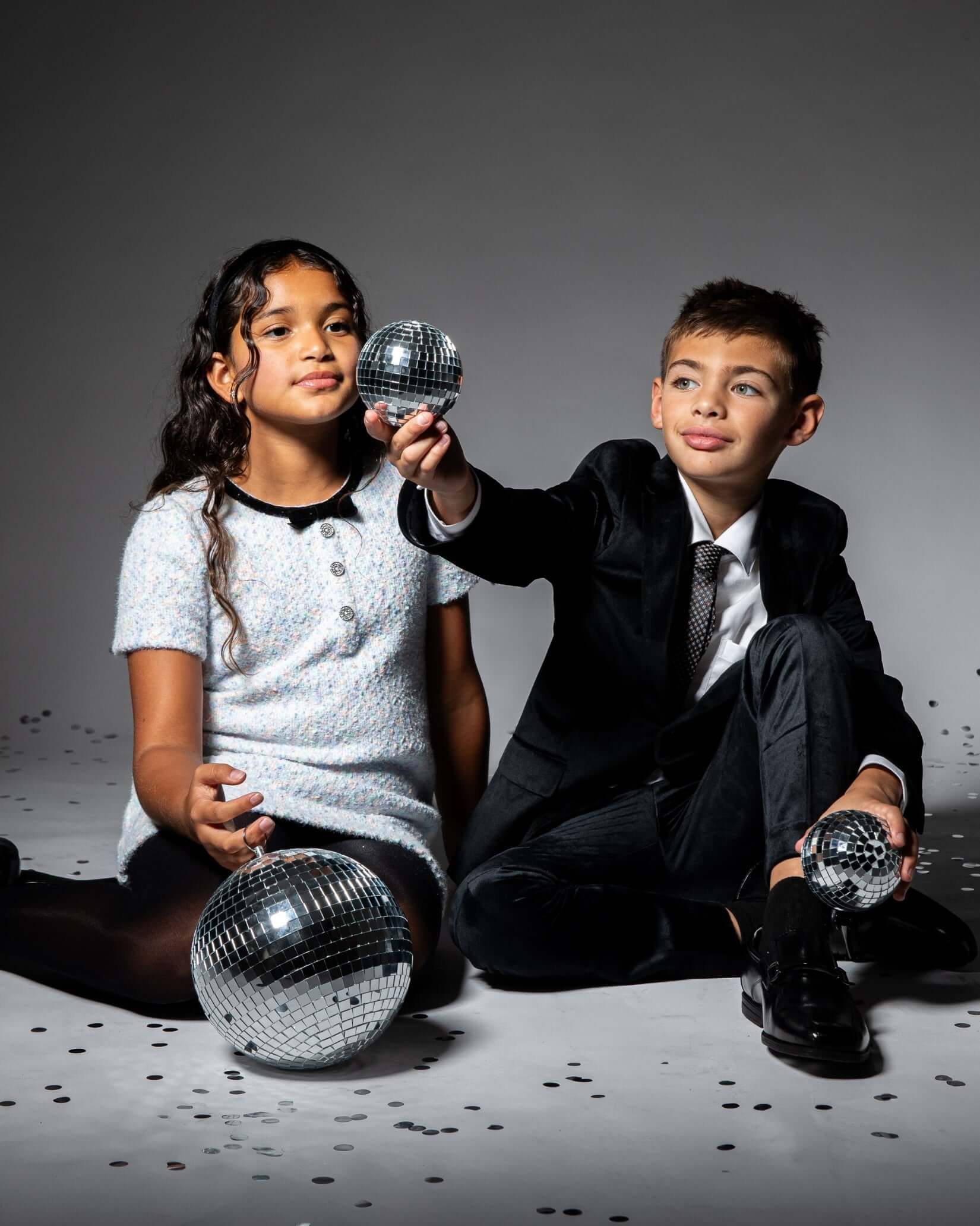 Two children sitting with disco balls, one wearing a dress and the other in a suit, ready for a festive celebration.