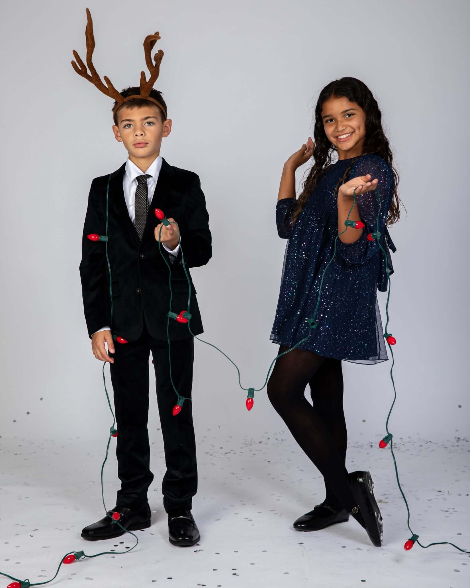 Two children dressed for a festive occasion, one in a suit with antlers and the other in a sparkly dress holding Christmas lights.