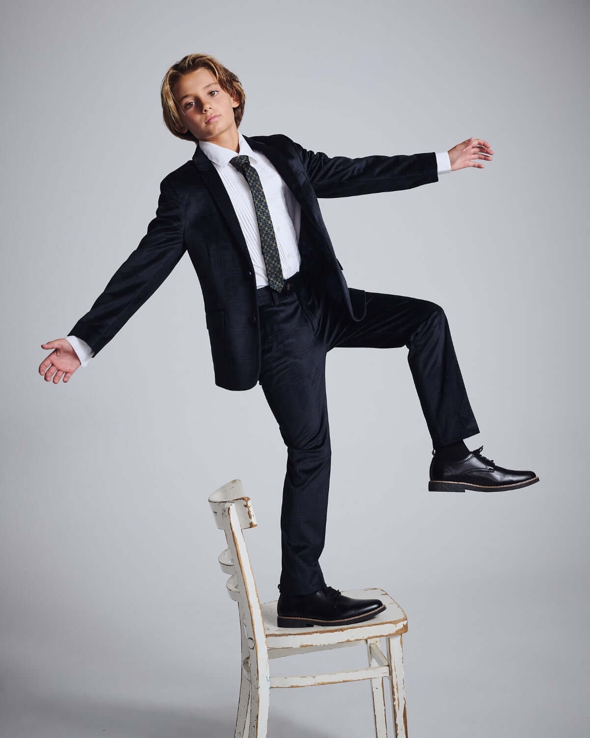 Boy modeling Midnight Velvet Plaid Suit Pants while balancing on a chair in a stylish pose.