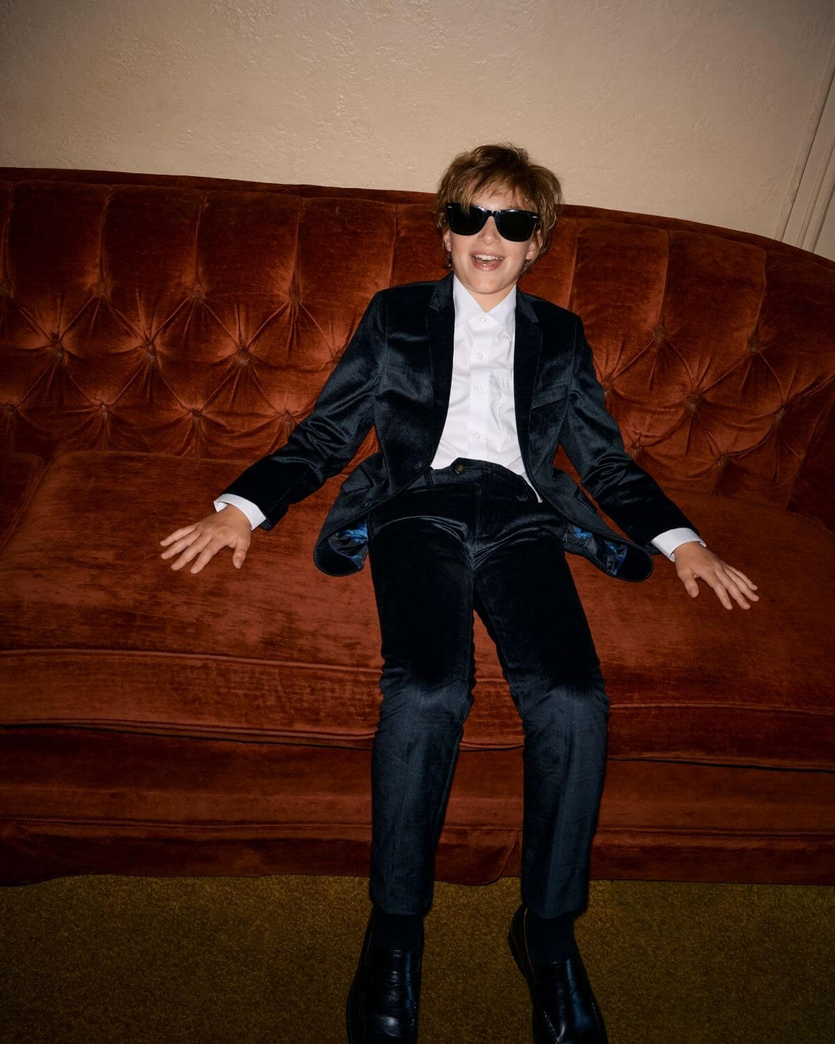 Child in stylish Midnight Velvet Plaid Suit Pants sitting on an orange velvet couch, wearing sunglasses.