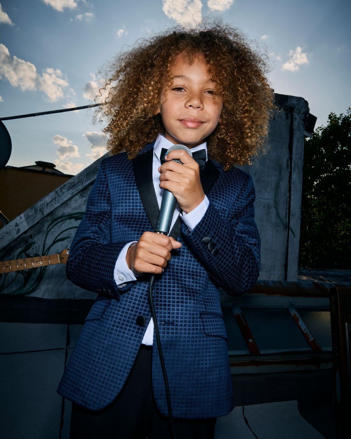 Boy wearing Navy Geo Velvet Tuxedo Jacket holding a microphone, exuding charm and confidence on a rooftop.