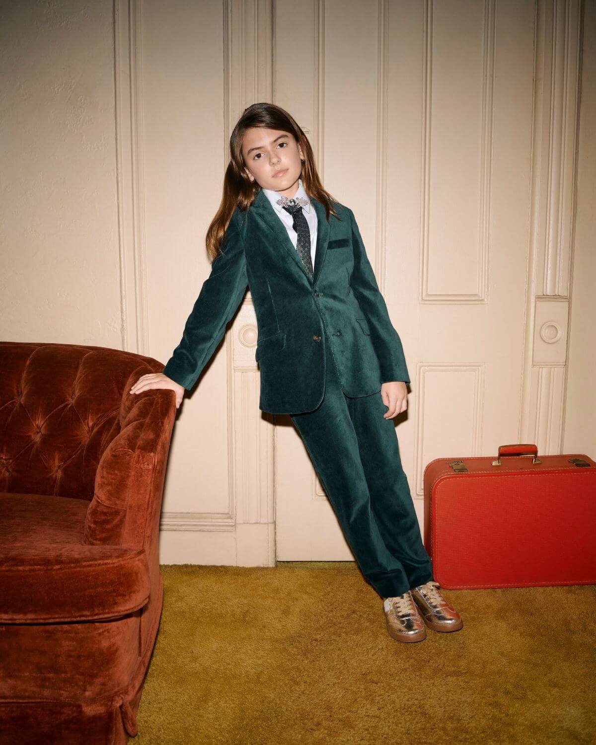 Evergreen Velvet Suit Pants on a child posing elegantly beside a vintage couch and a red suitcase.