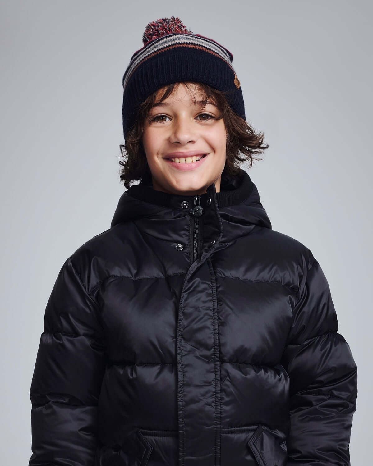 Child wearing a Navy Blue Wind Hat with a pom pom, smiling in a black puffer jacket.