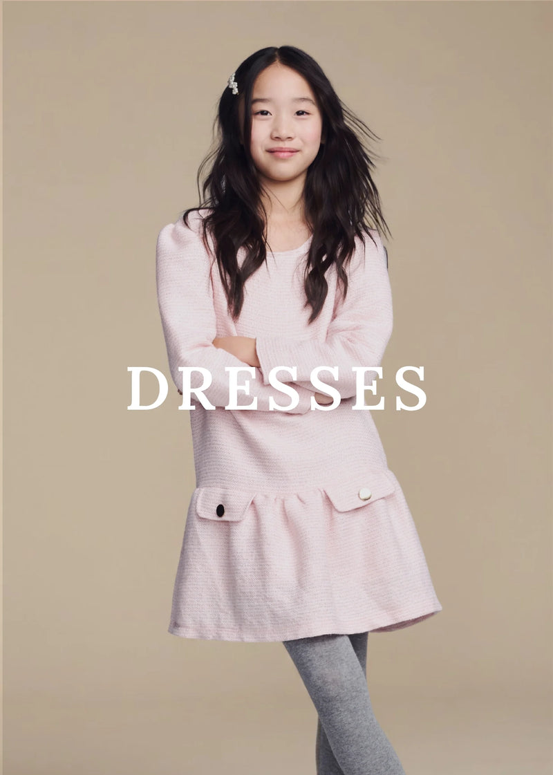 A young girl with long dark hair stands against a solid beige background wearing a light pink textured long-sleeve dress with grey leggings. The word 'DRESSES' is printed in white text across the center of the image.