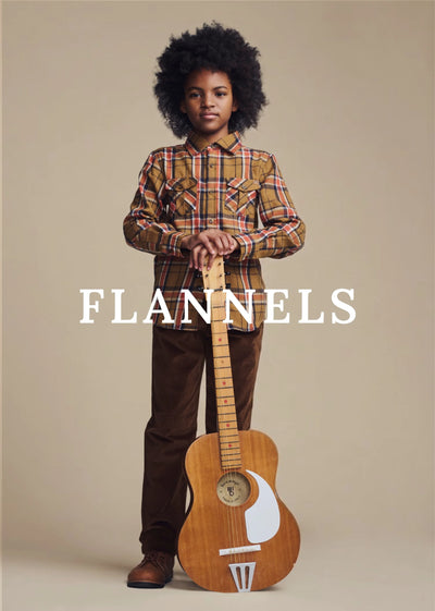 A child model with an afro hairstyle stands holding a wooden acoustic guitar, wearing a brown and red plaid button-down shirt and brown pants, against a solid beige background. The brand name 'FLANNELS' is printed in white text across the center.