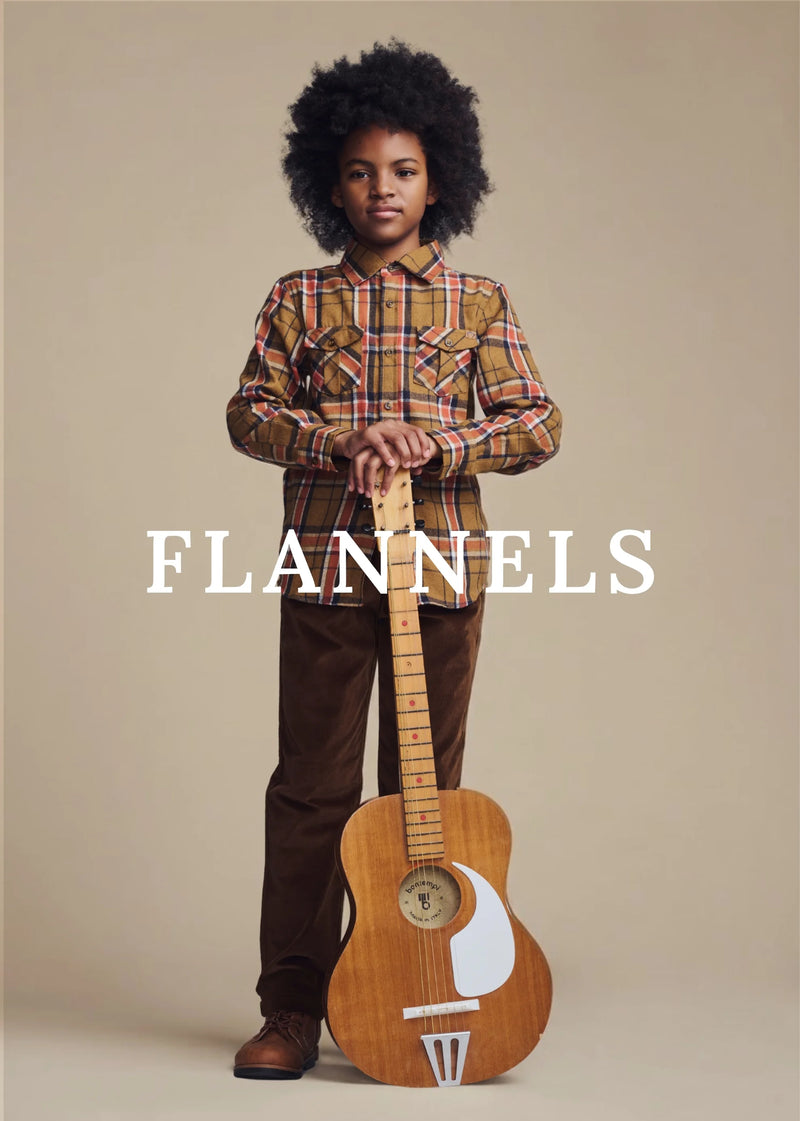 A child model with an afro hairstyle stands holding a wooden acoustic guitar, wearing a brown and red plaid button-down shirt and brown pants, against a solid beige background. The brand name 'FLANNELS' is printed in white text across the center.
