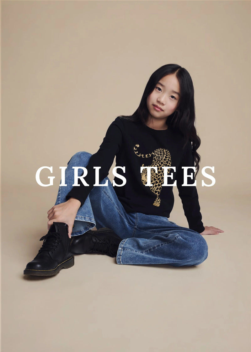 A young girl sits cross-legged on a beige studio background wearing a black long-sleeve graphic tee with a gold leopard print and blue jeans. Large white text across the center reads 'GIRLS TEES'.