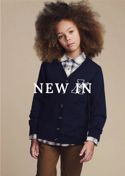 A young girl with voluminous curly brown hair is wearing a navy blue cardigan with a white embroidered letter 'A' logo, layered over a plaid button-down shirt and brown pants, set against a solid beige studio background with 'NEW IN' text overlay.