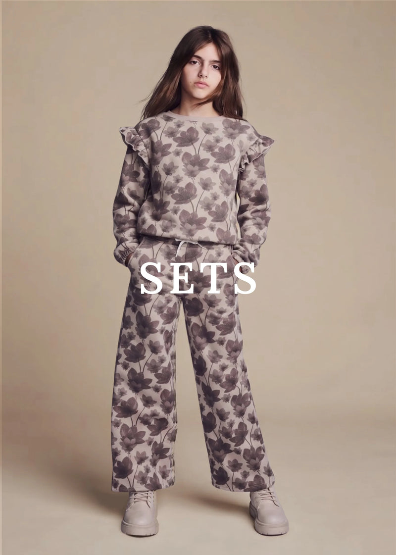 A young girl stands against a solid beige studio background wearing a matching two-piece tracksuit with a dark gray floral pattern on a light taupe base. The set includes a long-sleeve top with ruffled shoulders and matching wide-leg pants.