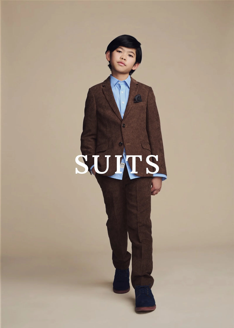 A young boy modeling a brown corduroy suit with a light blue dress shirt and a blue tie, standing against a solid beige studio background. The word 'SUITS' is printed in white text across the center of the image.