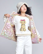 Girl wearing a Cream Bianca Graphic Sweatshirt with a cute bear graphic, styled with a shiny pastel jacket.