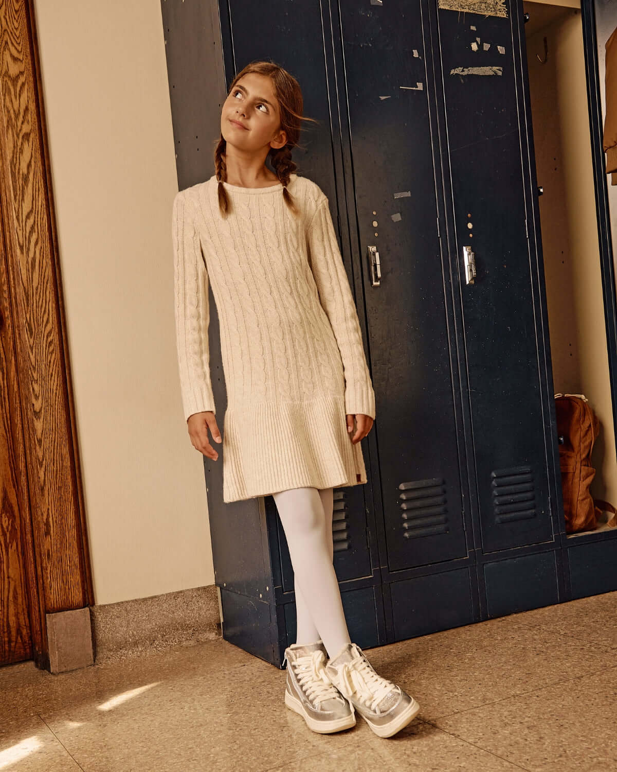 Young girl wearing a Cream Fisherman Sweater Dress with white leggings and stylish sneakers, standing in front of lockers.