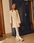 Young girl wearing a Cream Fisherman Sweater Dress with white leggings and stylish sneakers, standing in front of lockers.