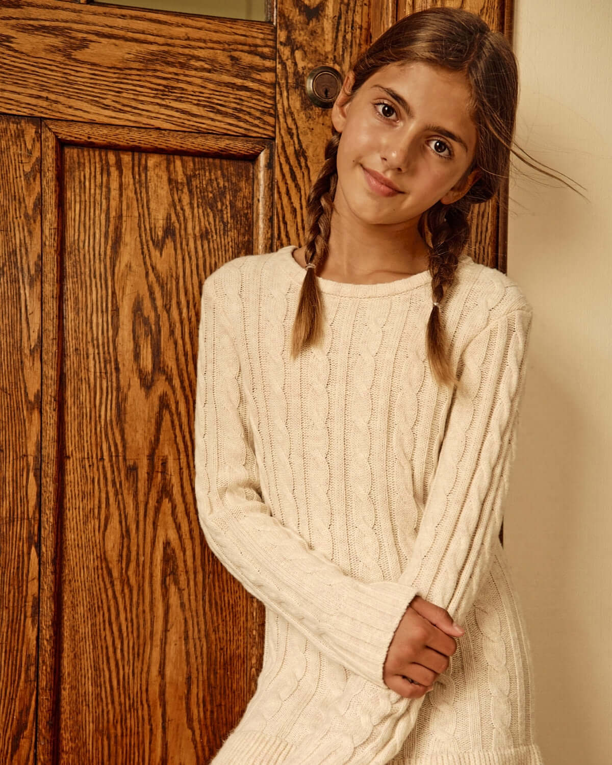 Girl wearing a Cream Fisherman Sweater Dress next to a wooden door, showcasing cozy knit style.