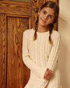 Girl wearing a Cream Fisherman Sweater Dress next to a wooden door, showcasing cozy knit style.