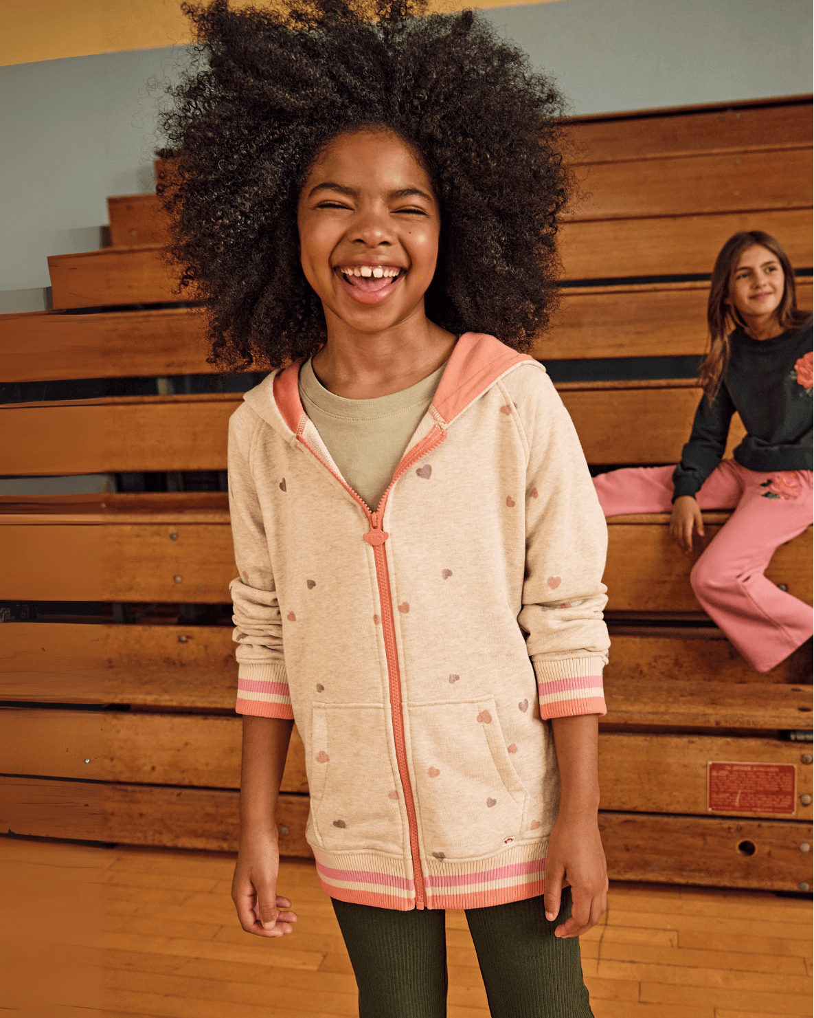 Heart Oatmeal Cozy Raglan Hoodie worn by a girl smiling in a gym setting, featuring pink and orange heart patterns.