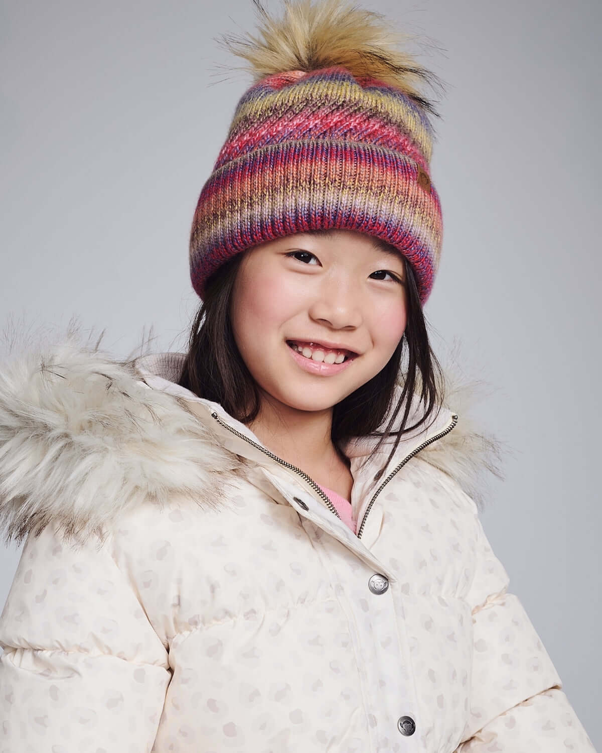 A smiling girl wearing a Rose Iris Pom Beanie with a cozy winter jacket, showcasing warm and stylish winter fashion.