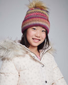 A smiling girl wearing a Rose Iris Pom Beanie with a cozy winter jacket, showcasing warm and stylish winter fashion.