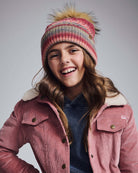 Happy girl wearing a Rose Iris Pom Beanie with a playful pom pom, styled in pink, red, and beige tones.
