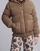 A cropped view of a person wearing a brown sherpa fleece zip-front jacket with a hood and large front patch pockets, paired with floral pants, against a light gray studio background.