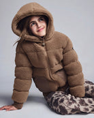 A young girl wearing a brown sherpa fleece hooded puffer jacket and patterned pants, sitting against a light gray studio background.