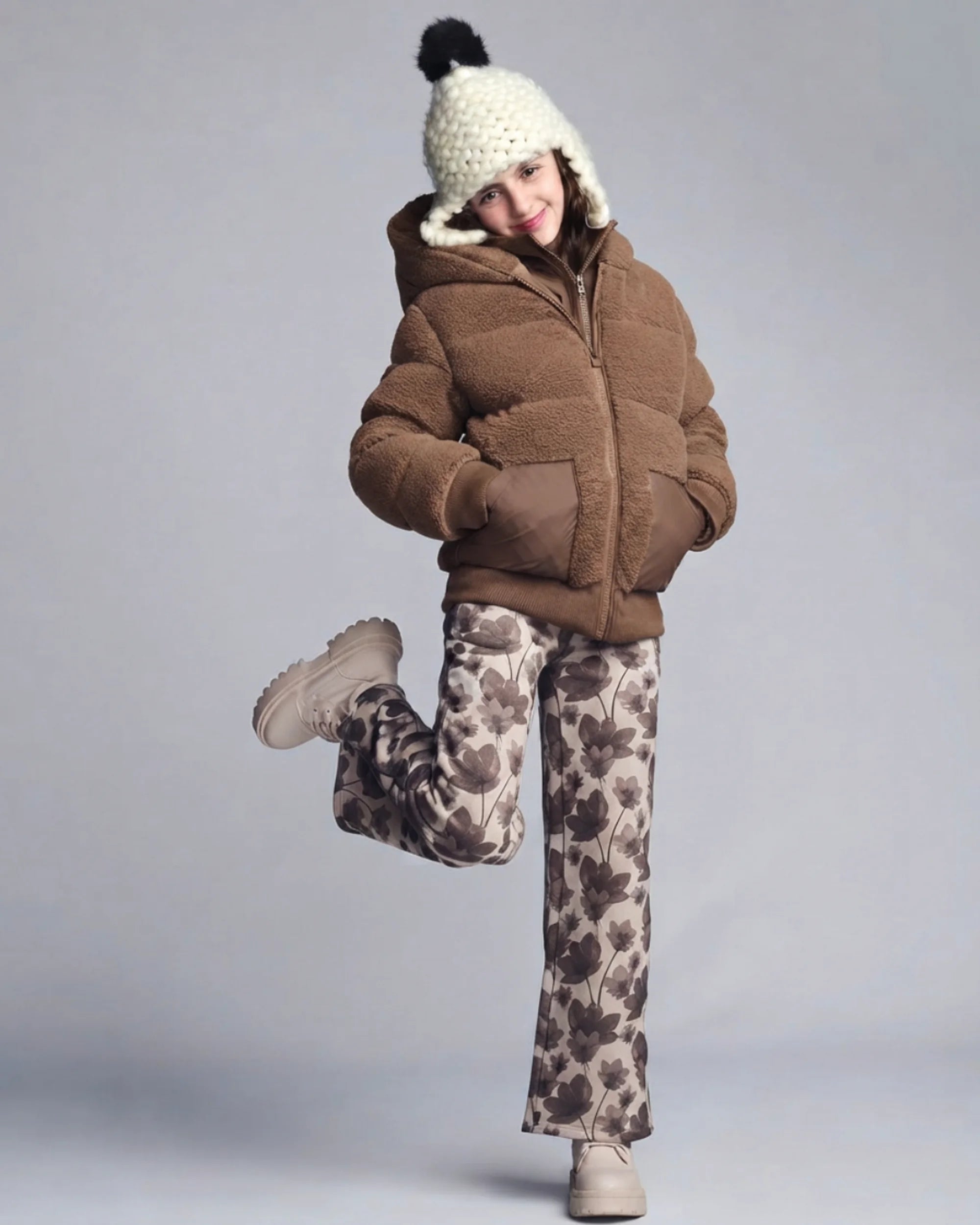 A child modeling a brown puffer jacket with a fuzzy sherpa-lined hood, paired with brown and beige floral print pants, a cream knit pom-pom hat, and beige boots, against a solid gray studio background.
