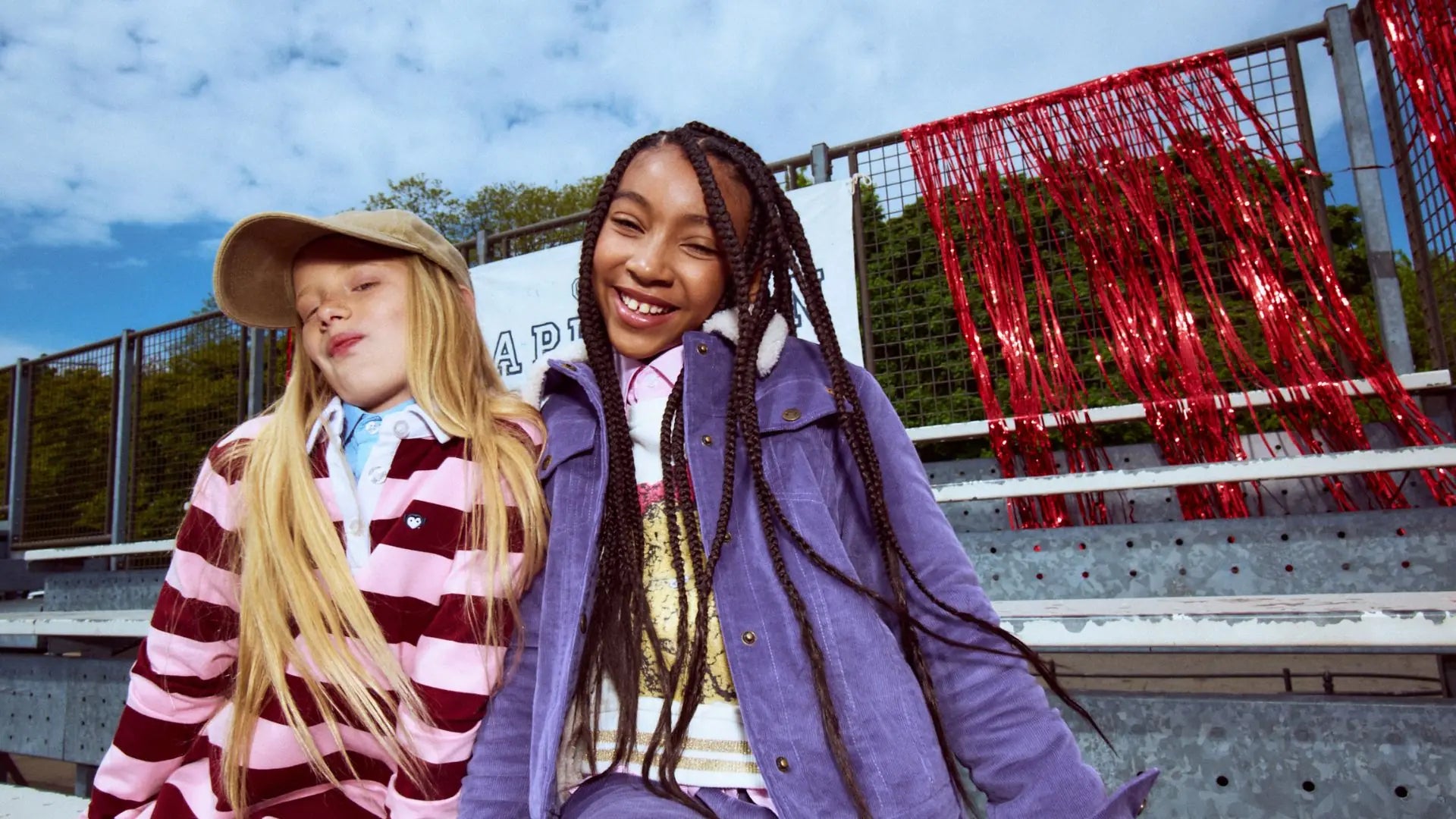 Two young girls sitting outdoors on stadium-style bleachers under a blue sky. The girl on the left wears a beige cap, a pink and burgundy striped jacket, and a light blue shirt.