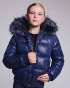 A young girl wearing a shiny navy blue puffer jacket with a dark faux-fur trimmed hood, standing against a light gray studio background.