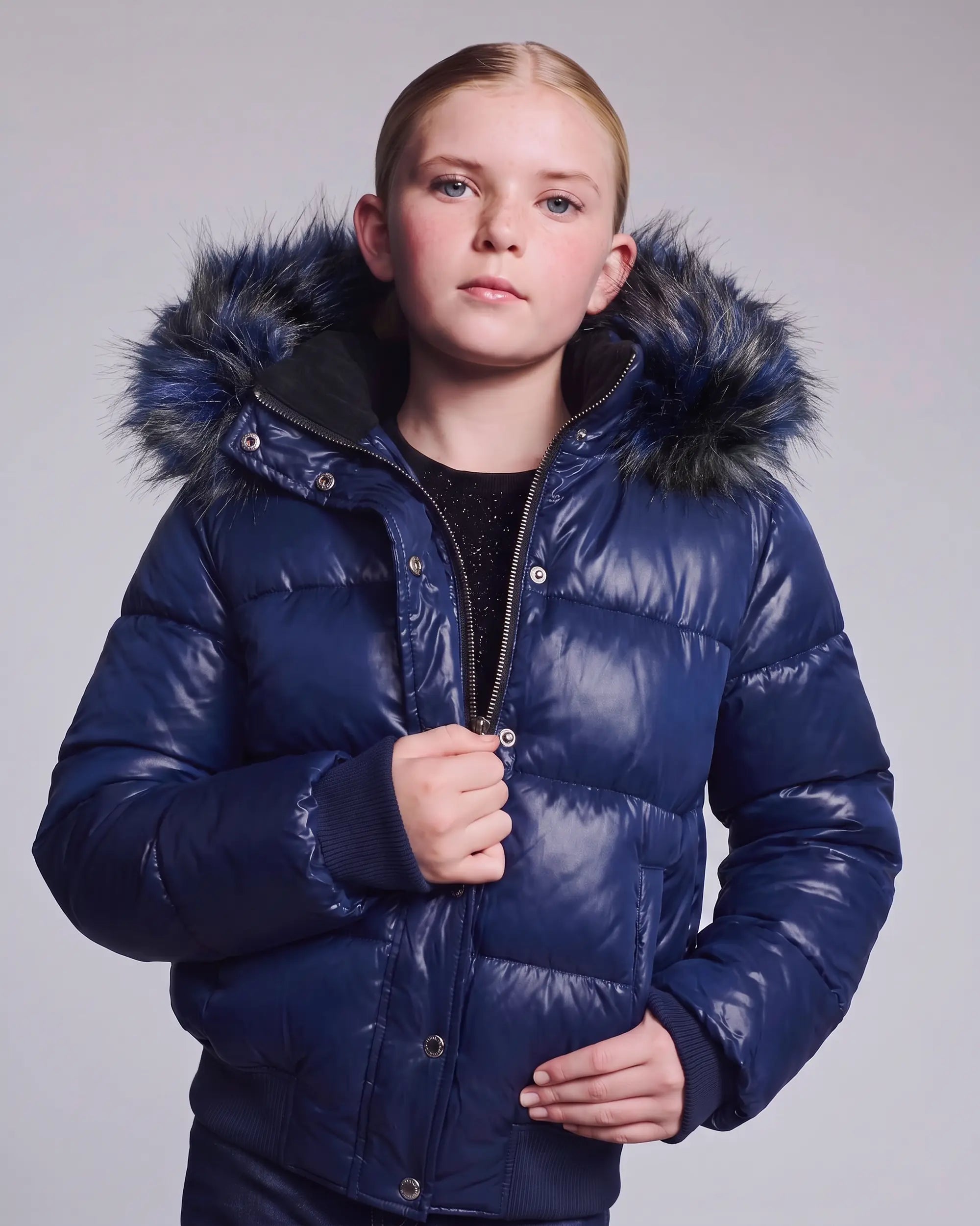 A young girl wearing a shiny navy blue puffer jacket with a dark faux-fur trimmed hood, standing against a light gray studio background.