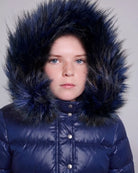 A close-up portrait of a child wearing a shiny navy blue puffer jacket with a large, fluffy dark navy faux-fur hood against a plain light gray background.