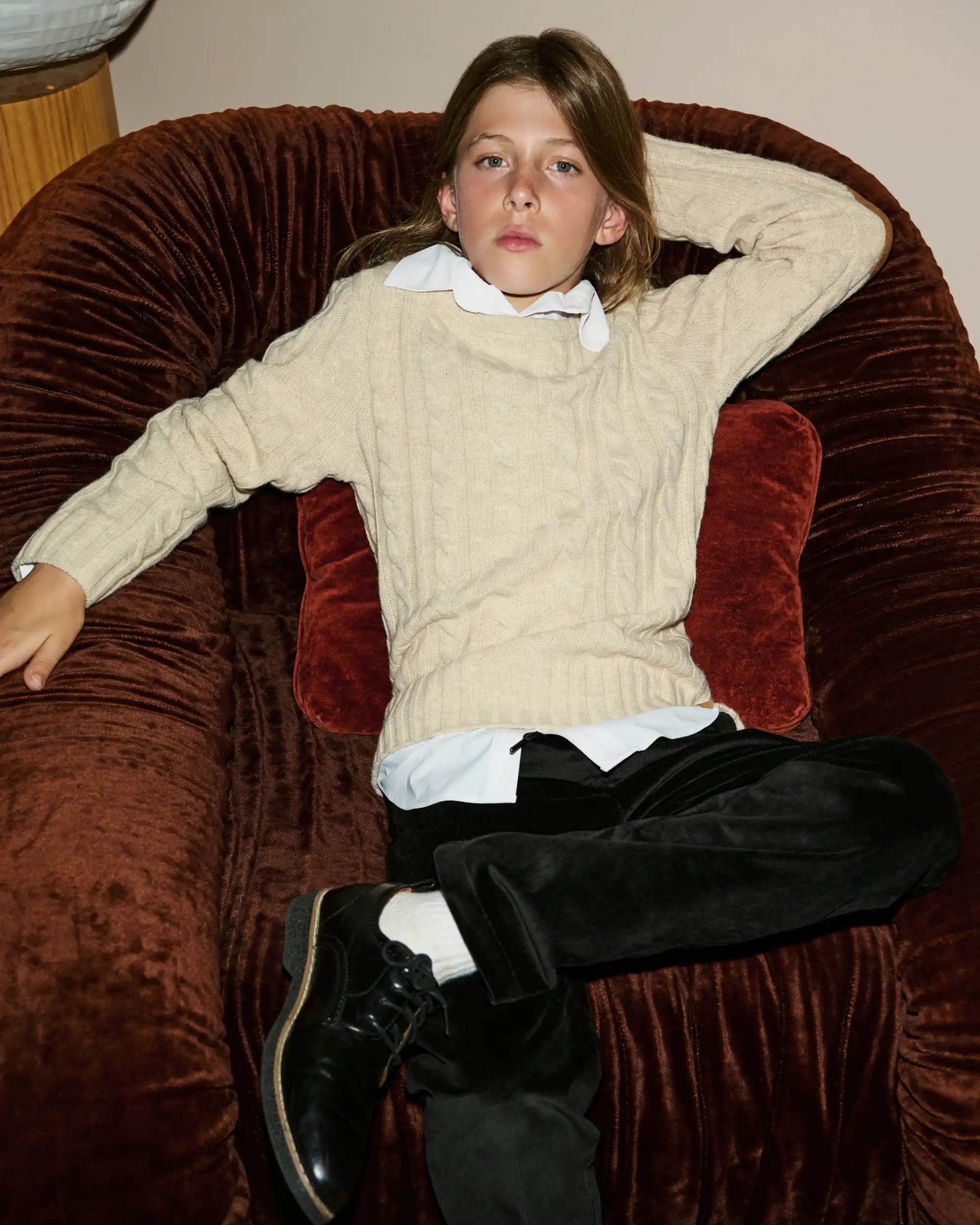 A young boy wearing a cream cable-knit sweater over a white collared shirt, black trousers, and black leather shoes, reclining on a brown velvet armchair.