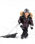 A happy child wearing a Burgundy/Black Kyla Puffer Coat playing in the snow and pulling a sled.