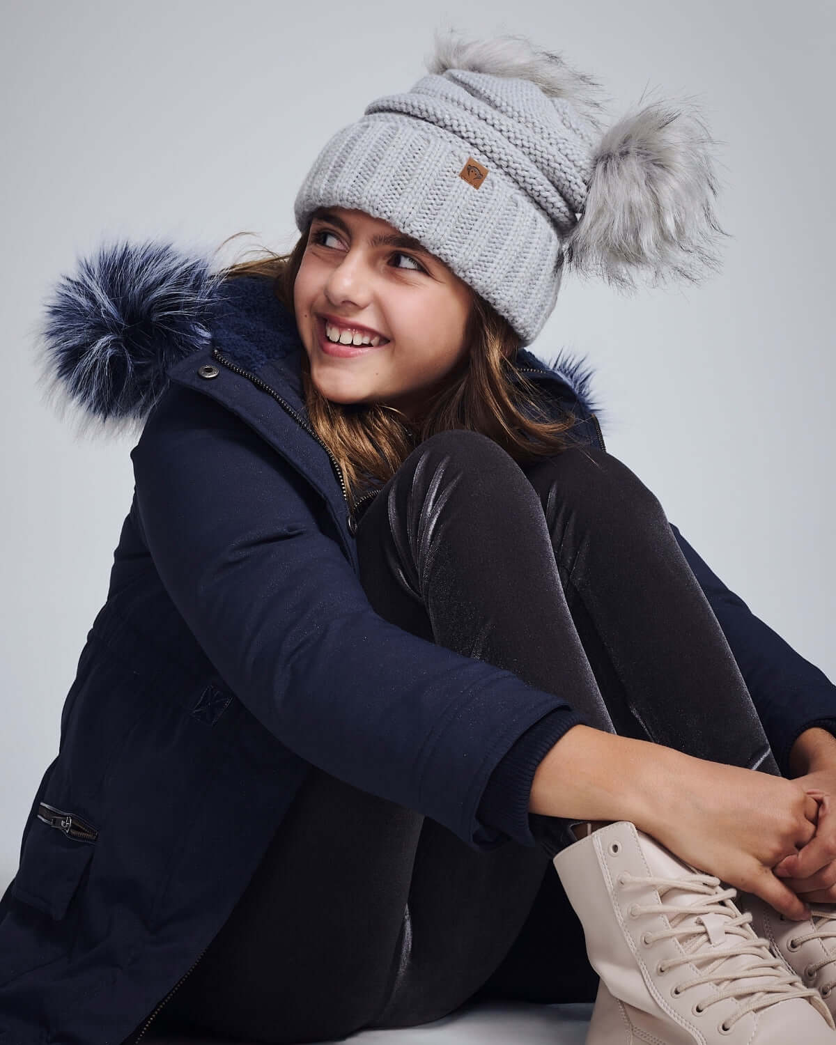 Girl wearing Shiny Silver Mia Faux Fur Pom Beanie with cozy winter coat and faux fur pom poms, smiling.