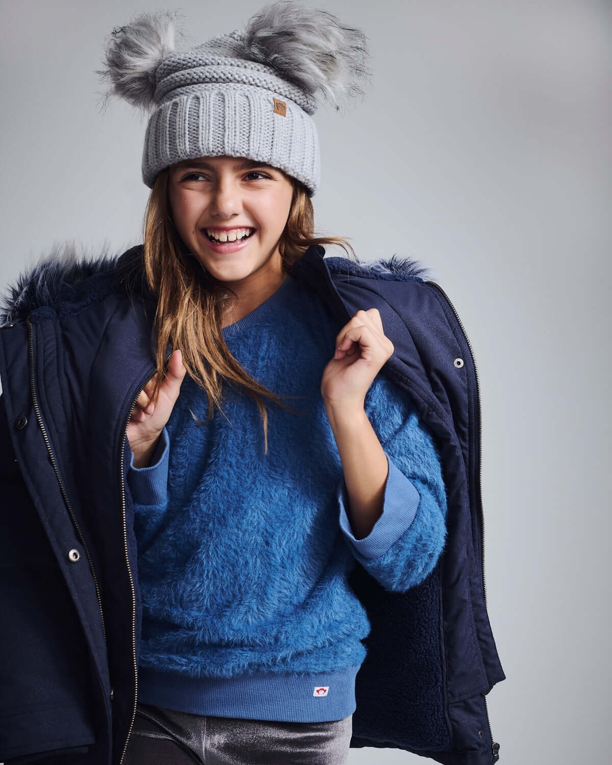 Shiny Silver Mia Faux Fur Pom Beanie worn by a girl in a blue fuzzy sweater and coat, smiling against a neutral background.