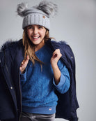 Shiny Silver Mia Faux Fur Pom Beanie worn by a girl in a blue fuzzy sweater and coat, smiling against a neutral background.