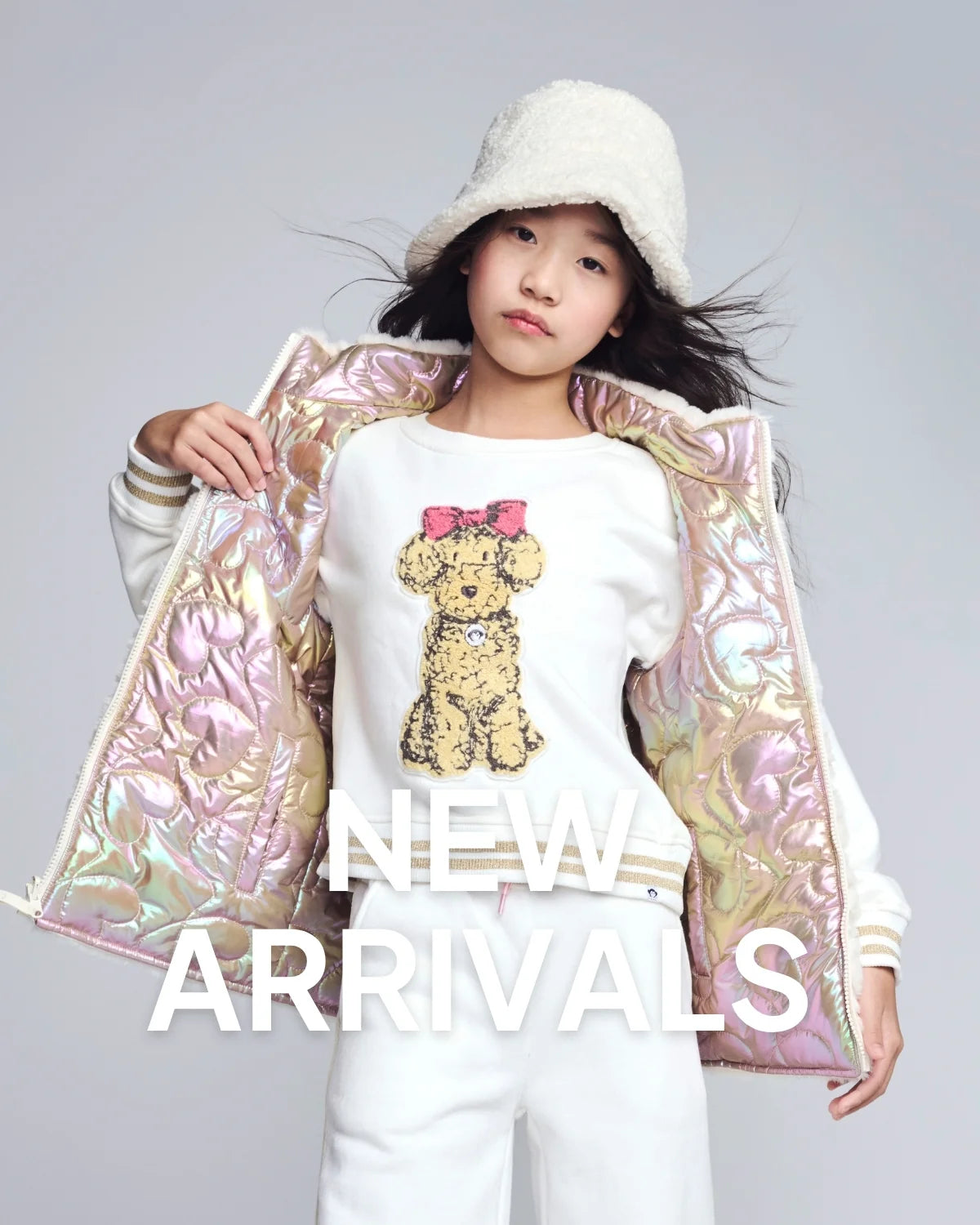 Child wearing a white outfit with a teddy bear design, holding a iridescent jacket, on a gray background with 'NEW ARRIVALS' text.