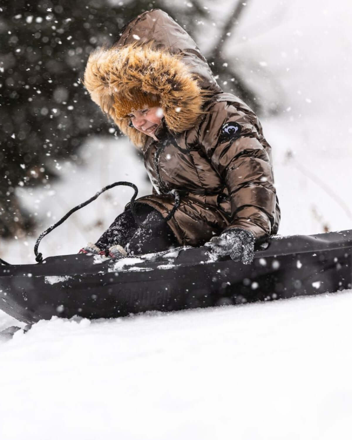 Girl wearing Copper Nova Long Coat while sledding in the snow, featuring faux fur hood and winter wonderland backdrop.