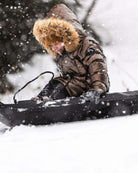 Girl wearing Copper Nova Long Coat while sledding in the snow, featuring faux fur hood and winter wonderland backdrop.