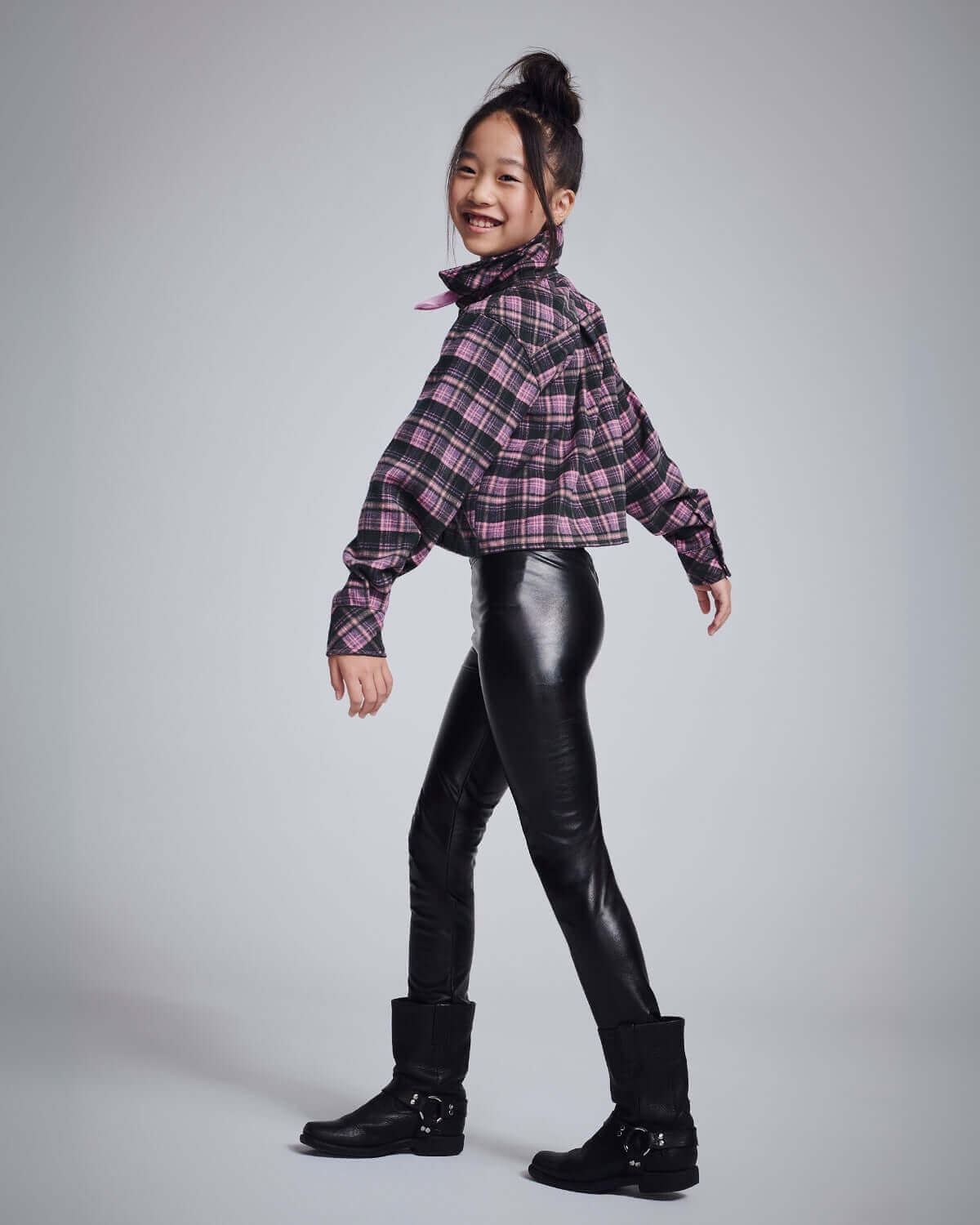 Girl wearing Pink Plaid Polly Cropped Flannel shirt with black leather leggings and boots, smiling playfully.